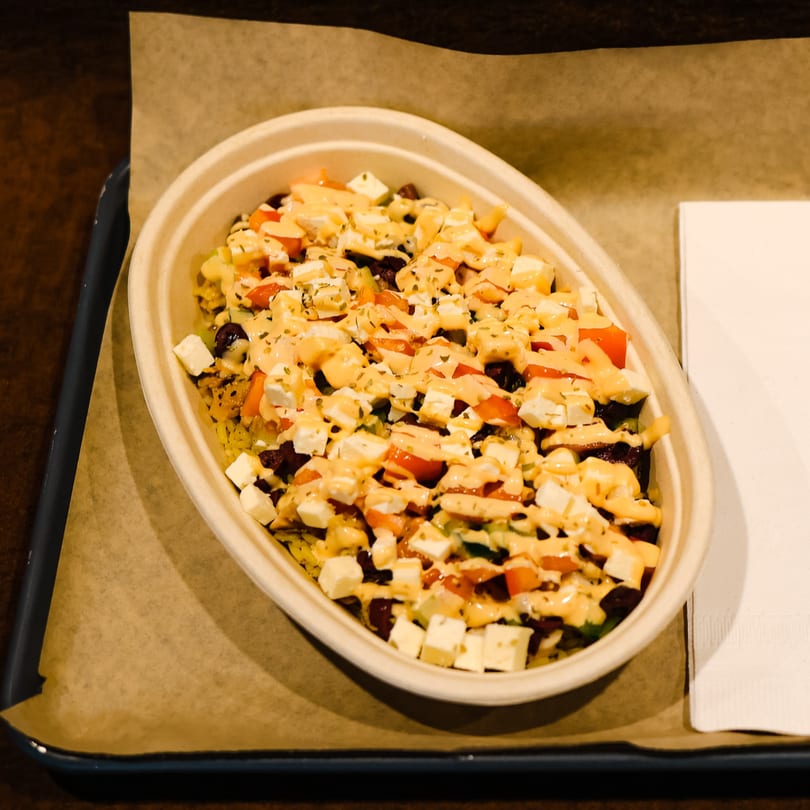A Greek bowl from Spyro's Gyros with feta, tomatoes, and sauce in a takeout container.