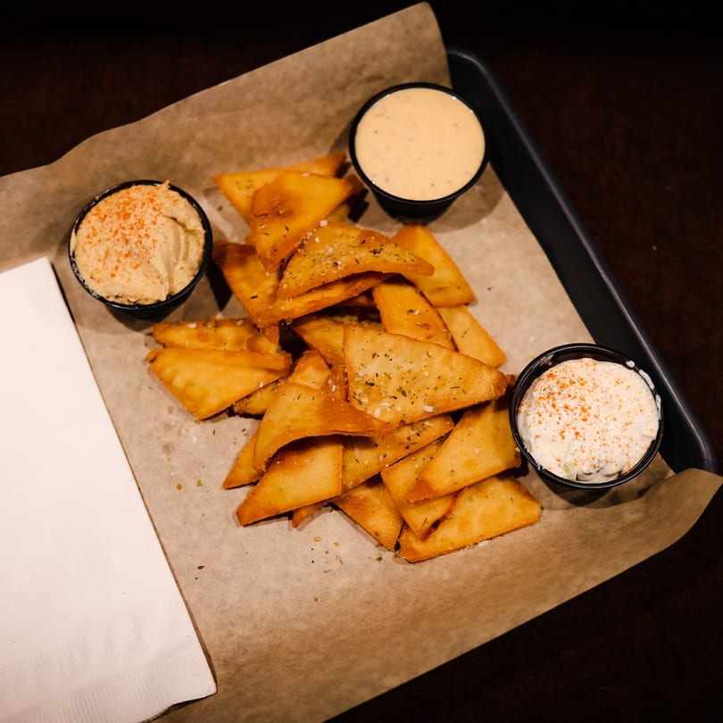 Crispy pita chips seasoned with herbs and salt, served with three dipping sauces on a tray.